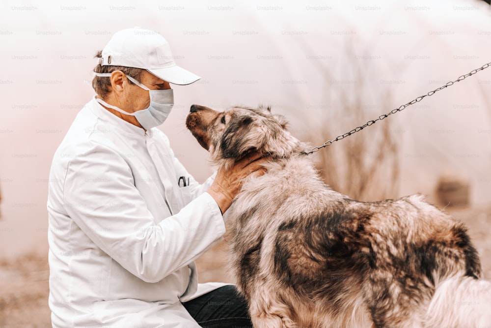 Veterinario maturo in camice bianco, maschera e berretto accovacciato e cane da accarezzare. Esterno rurale.