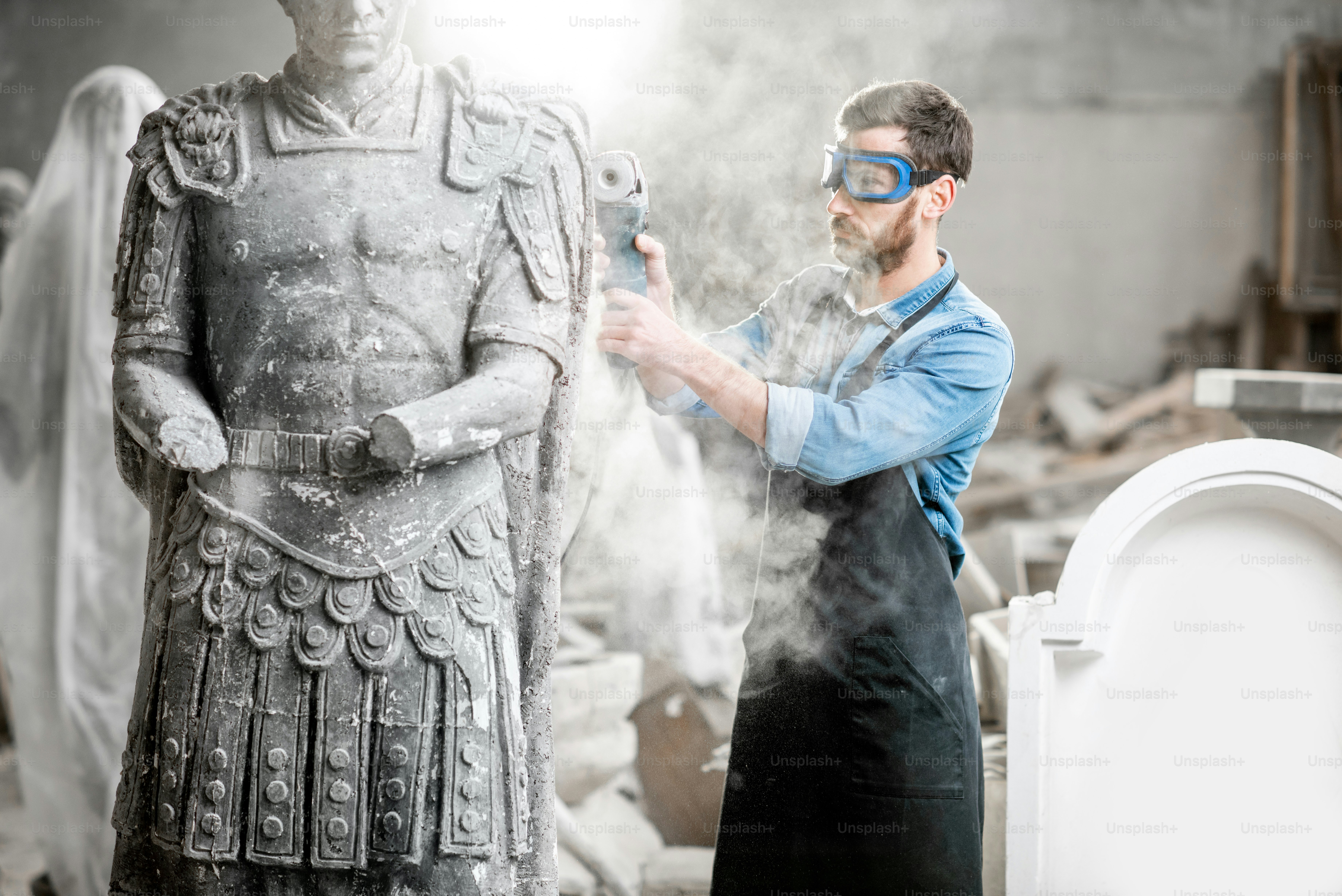 Sculptor in protective workware grinding stone sculpture with electirc grinder in the old studio with dust