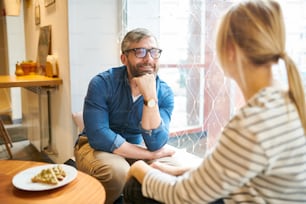 안경과 캐주얼웨어를 입은 행복한 수염을 기른 사랑스러운 남자가 아늑한 카페에서 대화하는 동안 여자 친구를 보고 있다