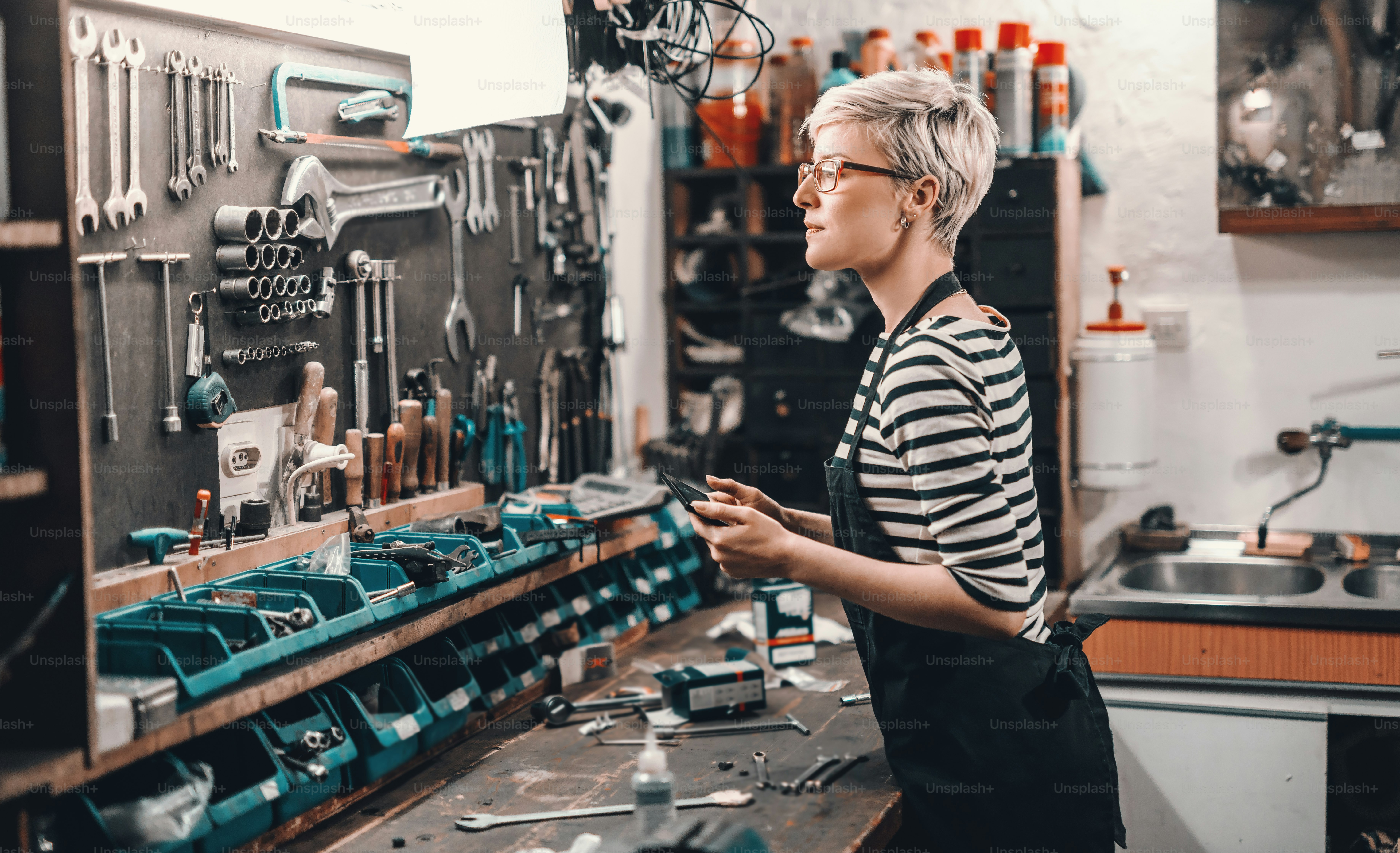 Bella operaia caucasica con capelli biondi corti e occhiali da vista che prendono attrezzo dal muro per riparare la bicicletta. Interno officina bici.