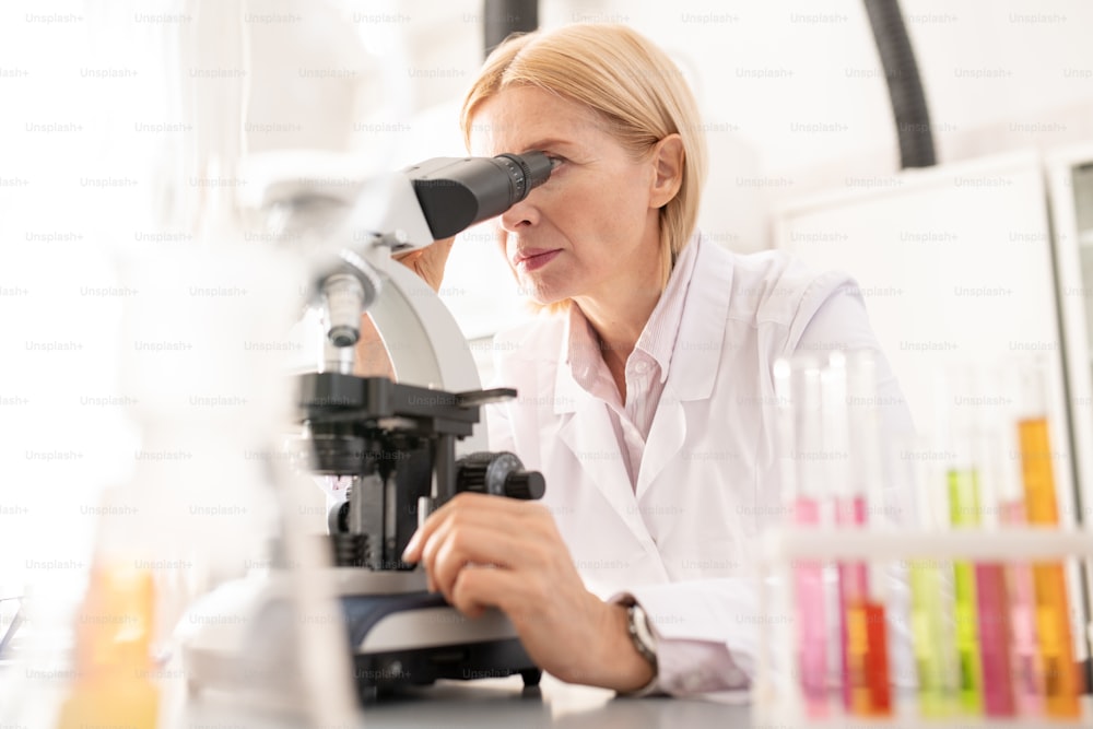 Serious concentrated mature lady in white coat working with microscope while doing scientific research in laboratory