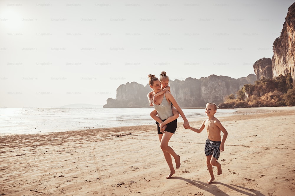 Laughing mother piggybacking her little daughter while running with her son along a sandy beach during summer vacation