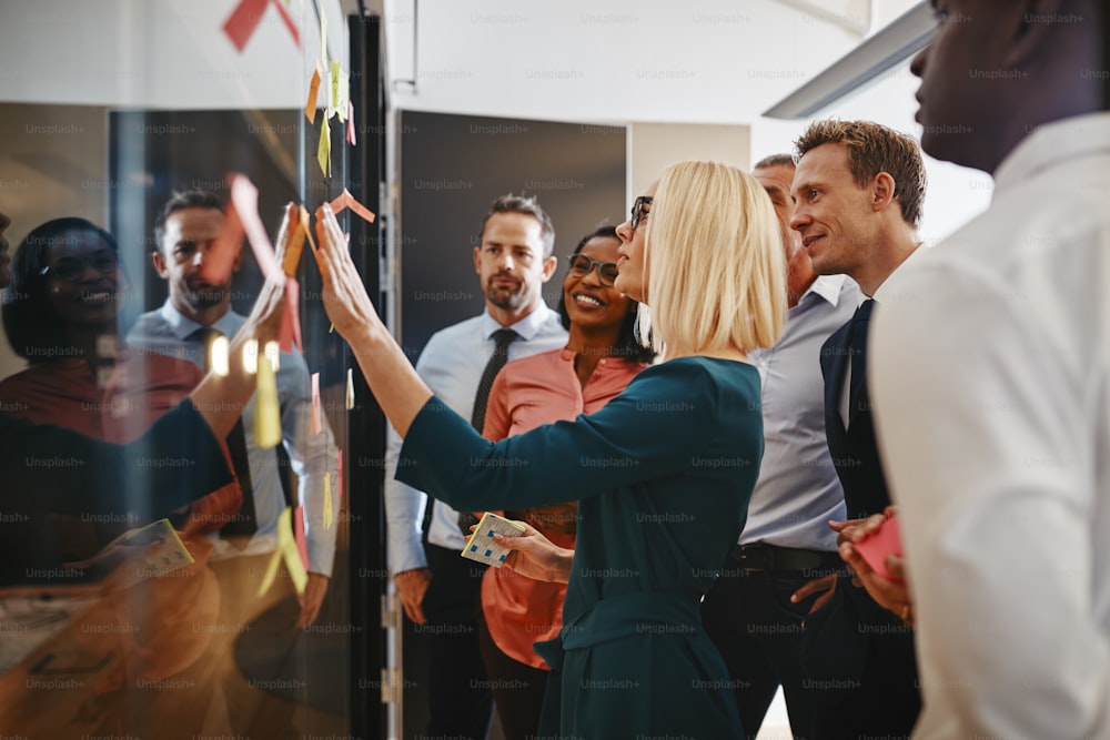 Une jeune femme d’affaires et son équipe diversifiée réfléchissent avec des notes autocollantes sur un mur de verre tout en travaillant ensemble dans un bureau moderne