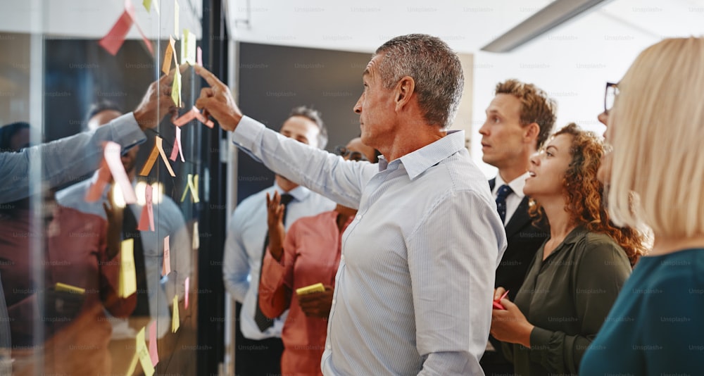 El gerente maduro y su equipo hacen una lluvia de ideas con notas adhesivas en una pared de vidrio mientras trabajan juntos en una oficina moderna