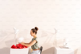 Vue de côté d’une jeune femme assise contre le mur près d’un bureau et cousant à la machine dans son propre studio