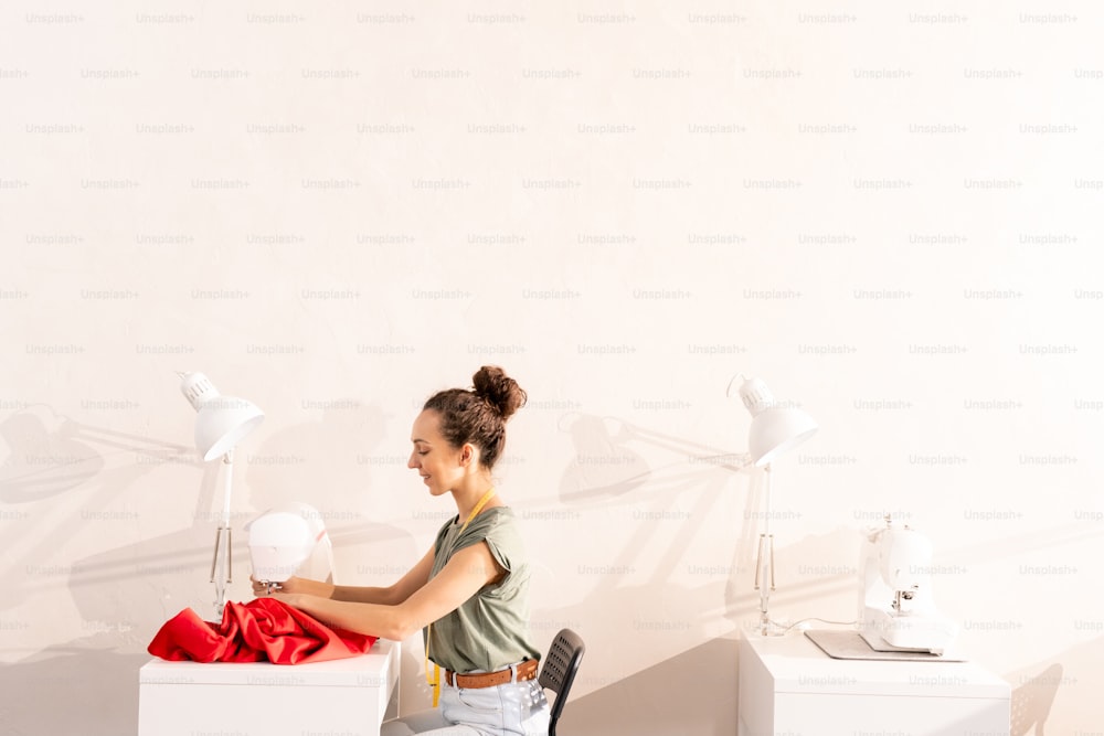 Side view of young female sitting against wall by desk and sewing by machine in her own studio