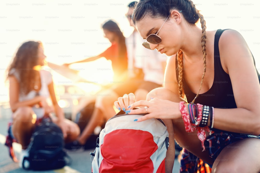 Serious Caucasian young woman opening backpack while crouching on the street. In background her friends chatting and looking at map. Summer holiday concept.