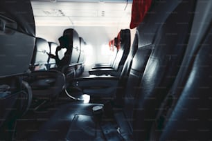 Wide-angle view of the row of seats inside of a modern aircraft with portholes at the end, dark vehicle interior with red rags under head and leather navy blue seats with belts on them