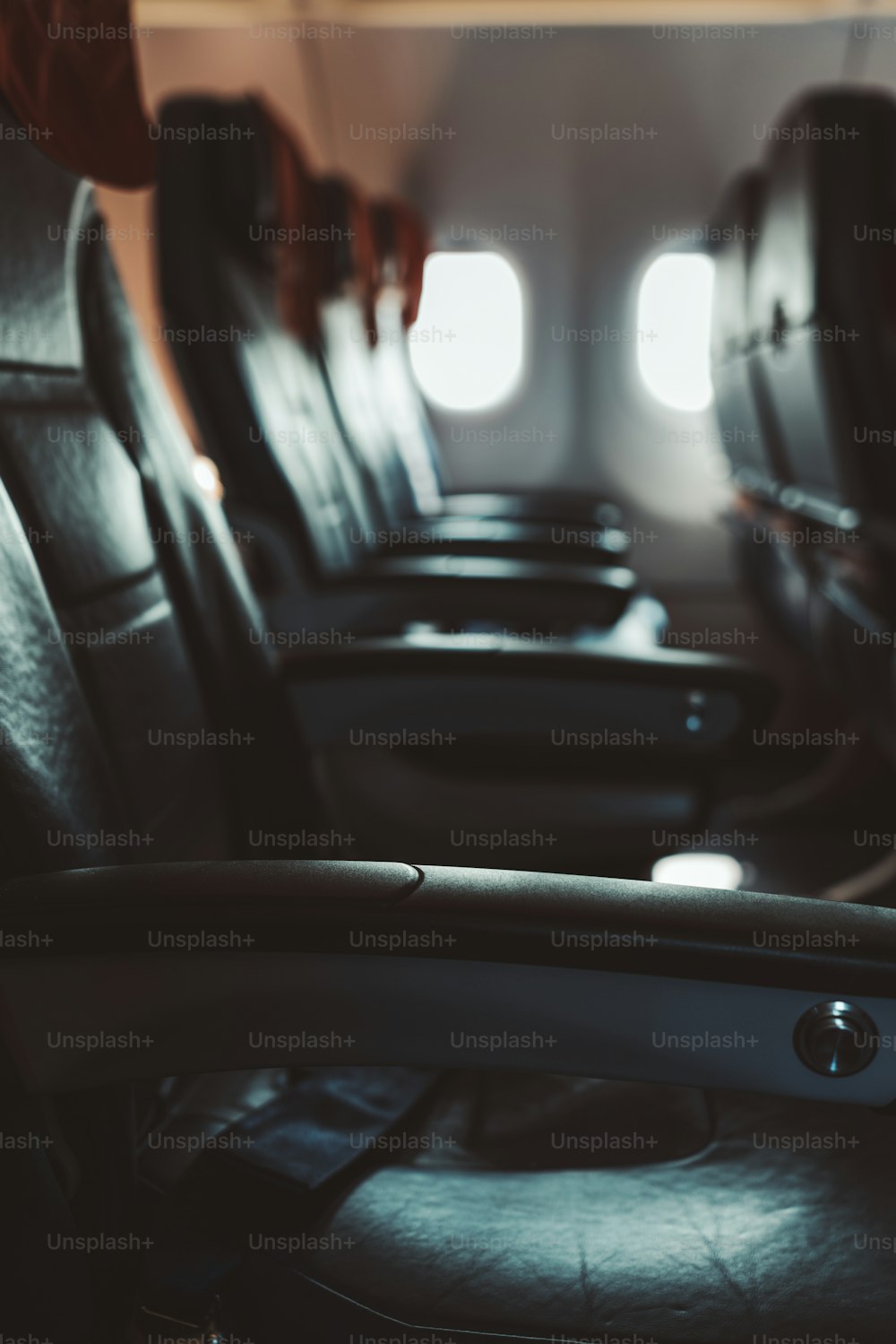 Un interior oscuro de avión en clase económica: la fila de modernos asientos de cuero vacíos con reposabrazos hacia abajo, poca profundidad de campo, enfoque selectivo en el reposacodos en primer plano, alfombra de trapo debajo de la cabeza