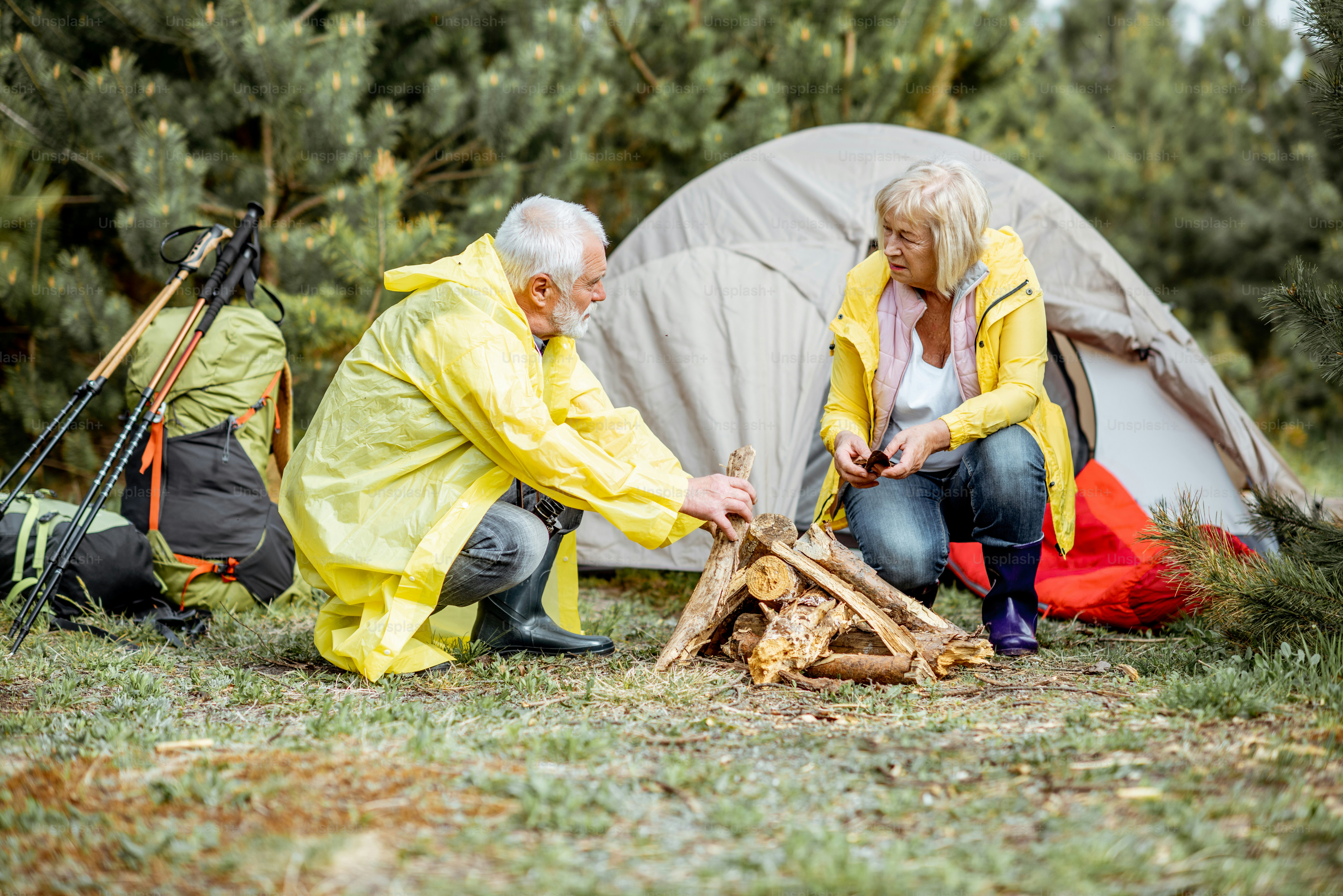 Seniorenpaar in gelben Regenmänteln beim Kamin auf dem Campingplatz in der Nähe des Zeltes im Wald
