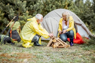 Seniorenpaar in gelben Regenmänteln beim Kamin auf dem Campingplatz in der Nähe des Zeltes im Wald