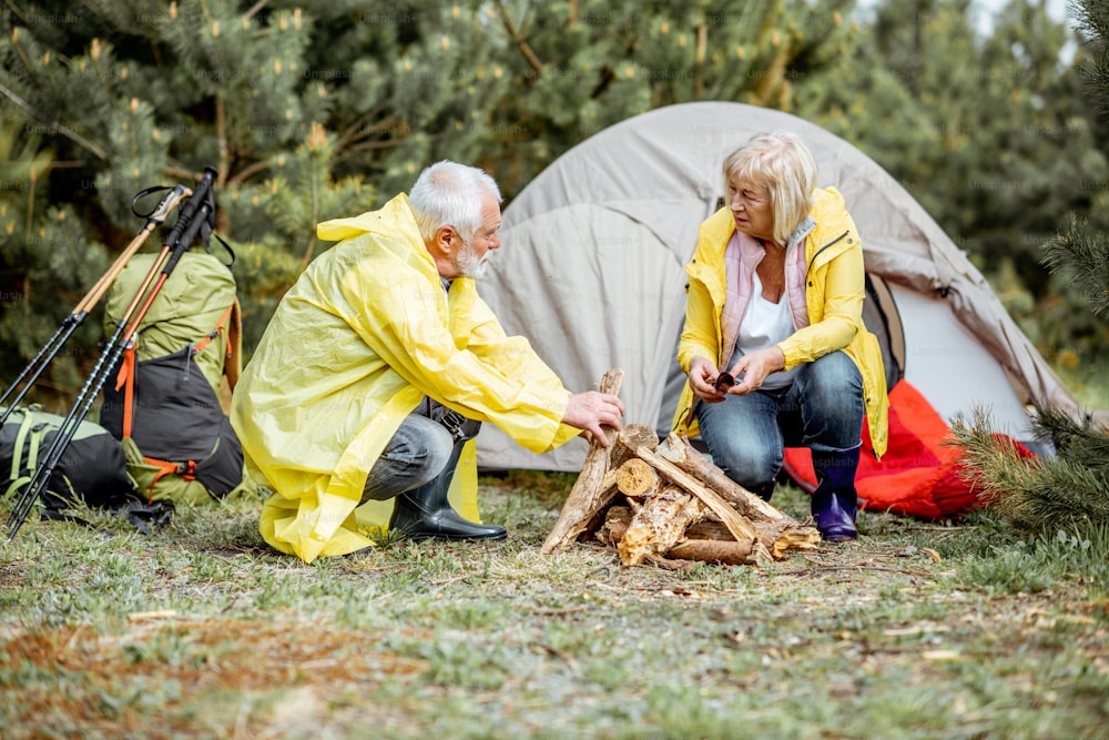Casal sénior em capas de chuva amarelas a fazer lareira no parque de campismo perto da tenda na floresta