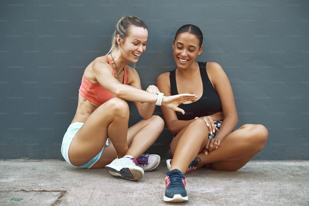 Fit young woman in sportswear sitting on the pavement outside showing her workout partner her sports watch while out for a run together