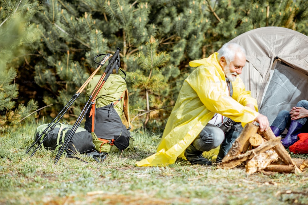 Senior Mann im gelben Regenmantel macht Kamin auf dem Campingplatz in der Nähe des Zeltes im Wald