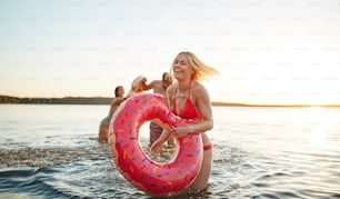 Laughing young woman in a bikini holding a swim ring with friends splashing in the background while having fun together at a lake