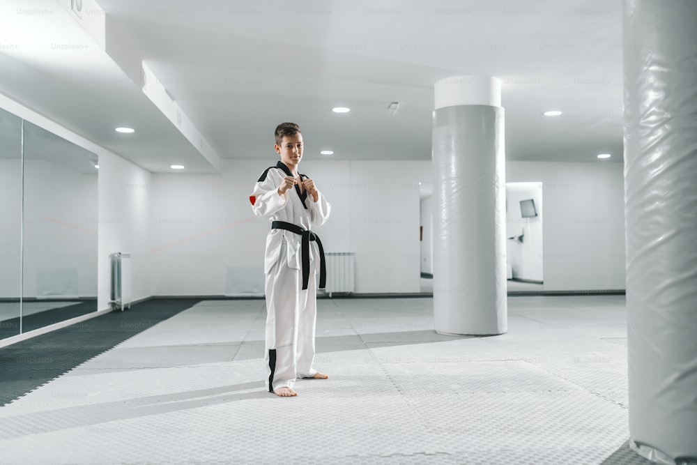 Boy in dobok standing barefoot, posing and looking at camera. Taekwondo training concept.