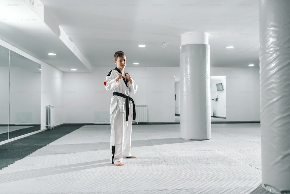 Boy in dobok standing barefoot, posing and looking at camera. Taekwondo training concept.