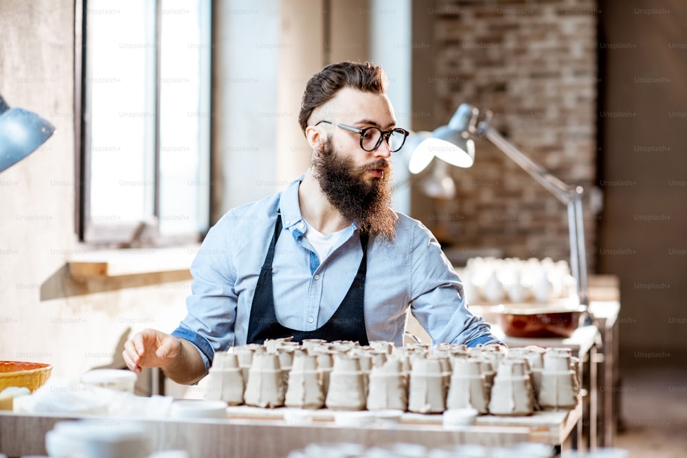 Handsome bearded man working with ceramic workpieces at the working place in the pottery shop