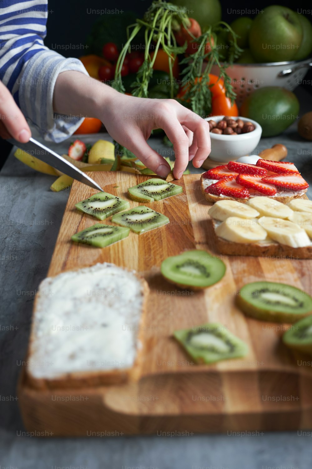 Close up of woman hands cutting fresh ingredients for sandwiches on the wooden board at the table surrounded by abundance of fruits