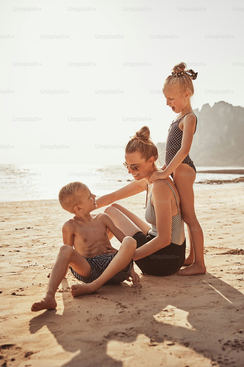 Madre risueña y sus dos adorables hijos sentados juntos en una playa de arena durante las vacaciones de verano