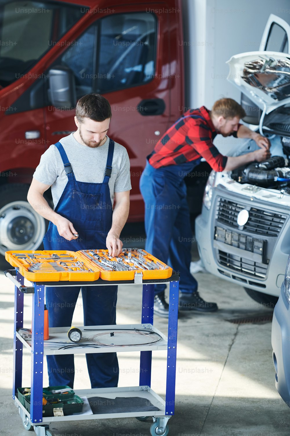 One of young technicians in workwear selecting handtools in toolbox on background of his colleague working