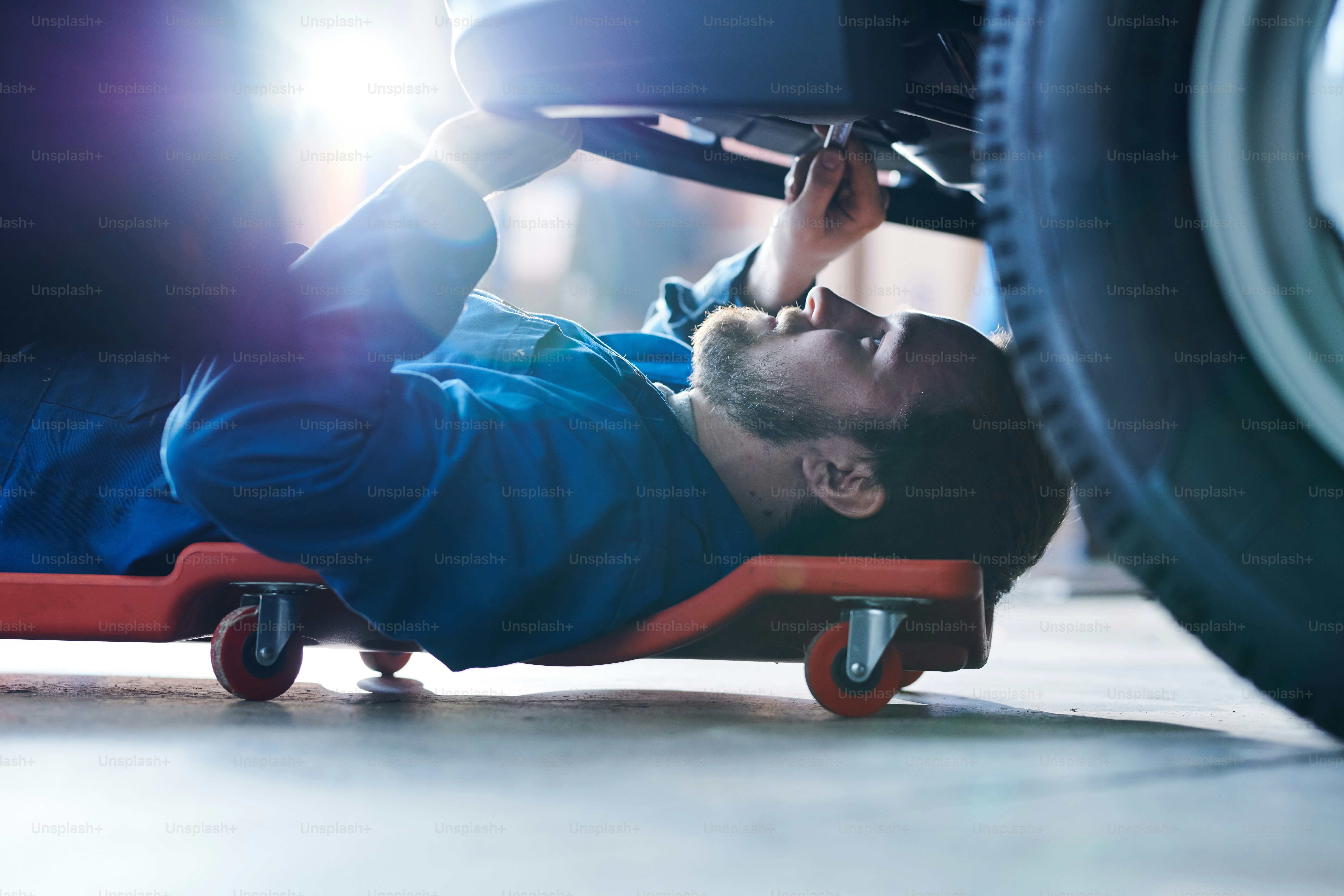Jovem trabalhador do centro de serviços de reparação de automóveis deitado no carrinho especial sob o automóvel e fixação de pequenos detalhes