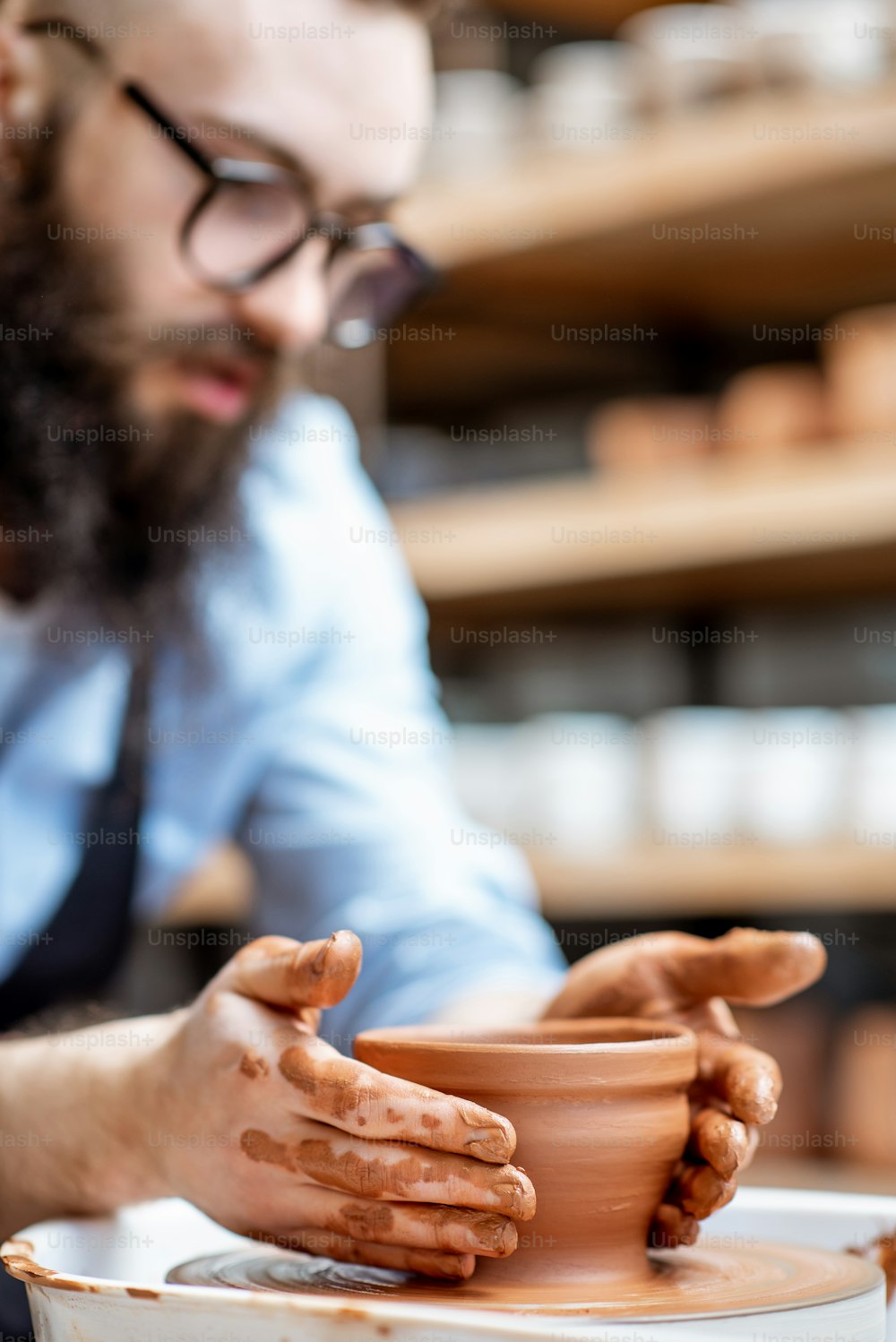 Hombre guapo como trabajador de alfarero en delantal haciendo jarras de arcilla en la rueda de cerámica en la pequeña fábrica