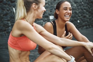 スポーツウェアを着た2人のフィット感のある若い女性の友人が、ランニング後に外の駐車場に座って一緒に話したり笑ったりしている