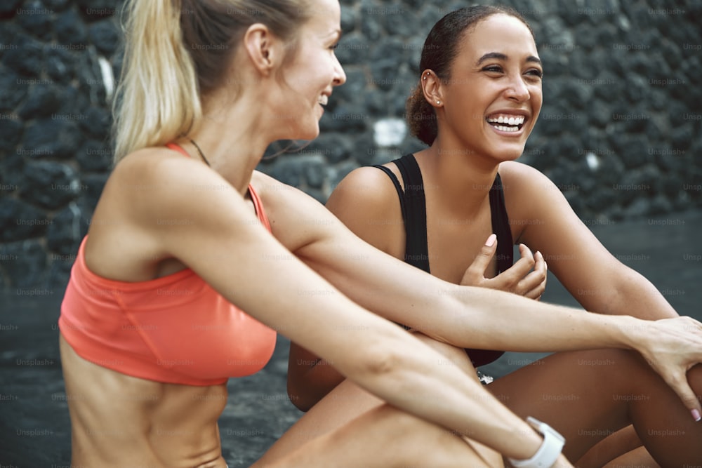 Zwei fitte junge Freundinnen in Sportbekleidung unterhalten und lachen miteinander, während sie nach einem Lauf draußen auf einem Parkplatz sitzen