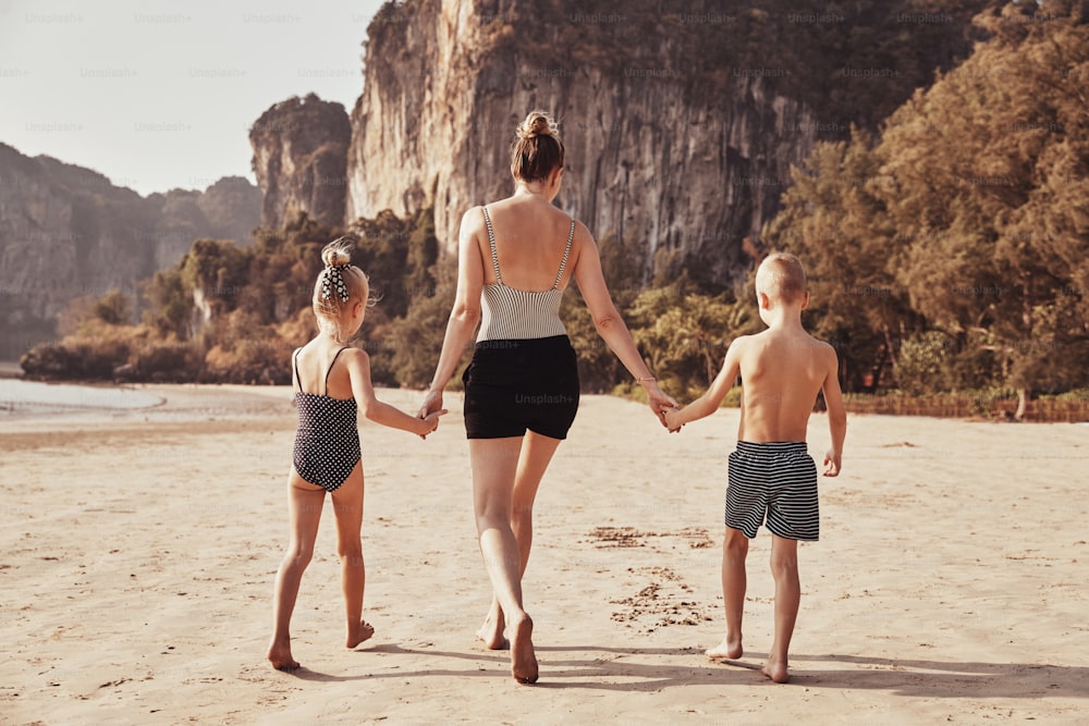 Vue arrière d’une mère et de ses deux enfants marchant main dans la main le long d’une plage de sable pendant les vacances d’été