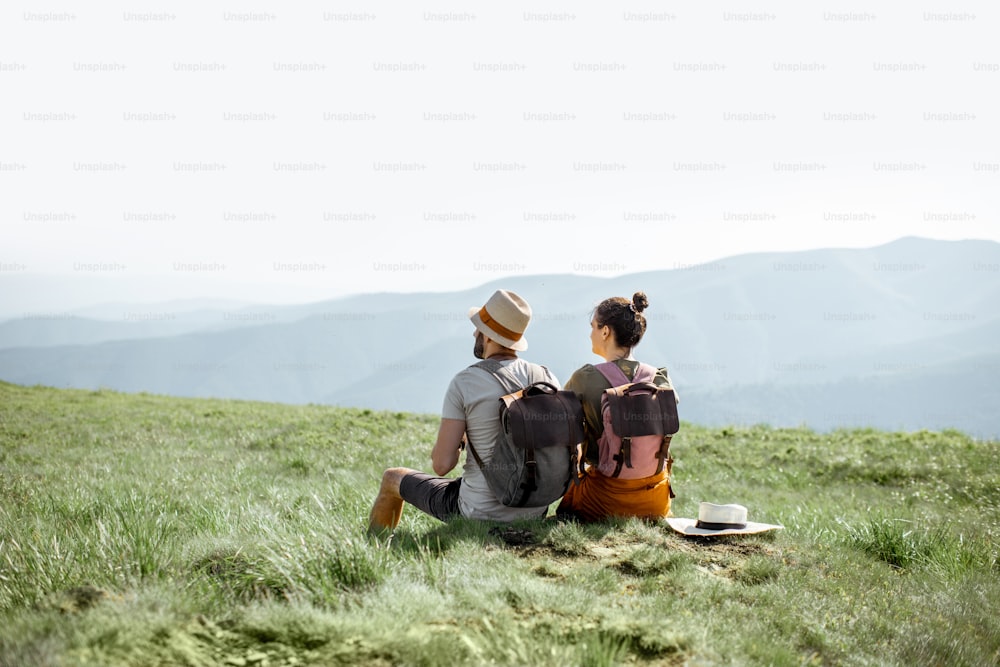 Giovane coppia seduta sul prato verde, che viaggia con zaini in montagna durante il periodo estivo