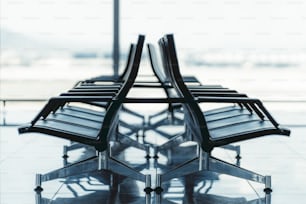 Blick auf zwei Reihen von Kunststoffsitzen im Innenbereich eines modernen Flughafenterminals oder eines Bahnhofsdepots mit großem Fenster dahinter, geringe Tiefenschärfe, selektive Fokussierung auf die nächstgelegenen Sessel