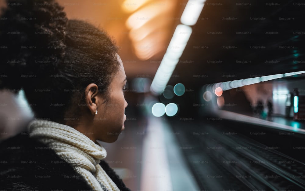 Echte Tilt-Shift-Seitenansicht einer jungen, schönen afroamerikanischen Frau, die auf den herannahenden Zug schaut, während sie auf dem Bahnsteig der U-Bahn-Station bleibt, mit einem Kopierplatz auf der rechten Seite für Ihre Anzeige
