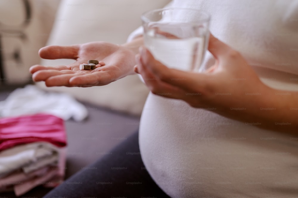 Close up of pregnant woman holding vitamins in one hand and glass of water in other.