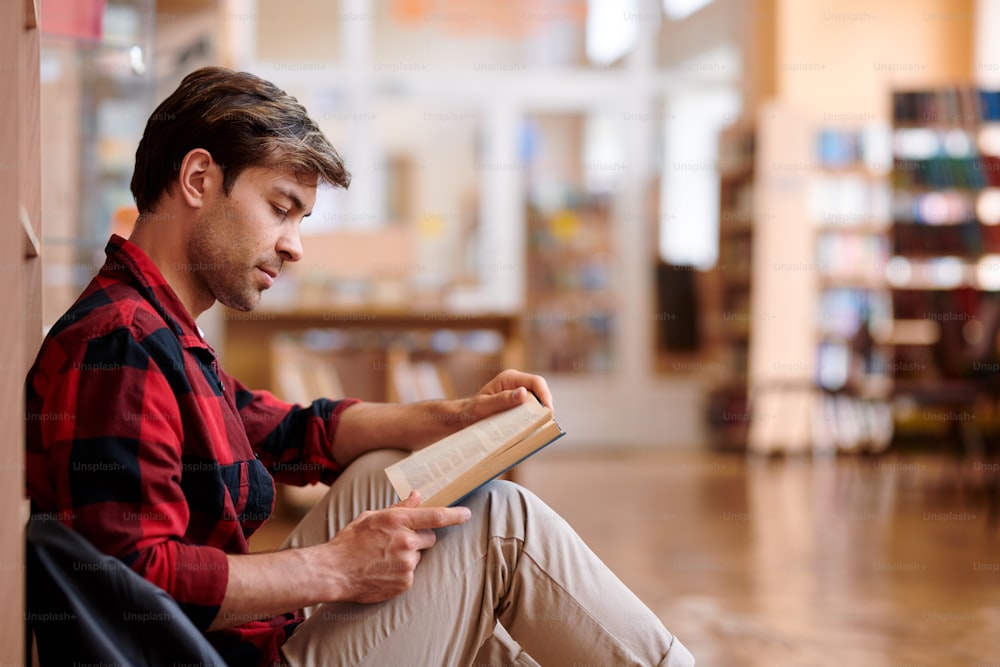 Lässiger junger Mann, der Buch liest, während er am Bücherregal in der Bibliothek des College sitzt