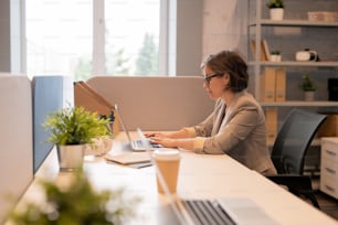 Serious concentrated office lady in glasses sitting at table and typing on laptop while preparing report in office