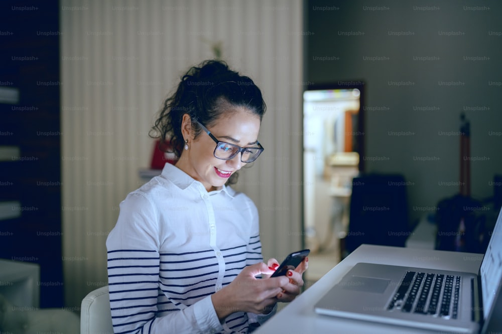 Gorgeous smiling businesswoman with brown curly hair, ponytail and eyeglasses sitting in office and using smart phone.