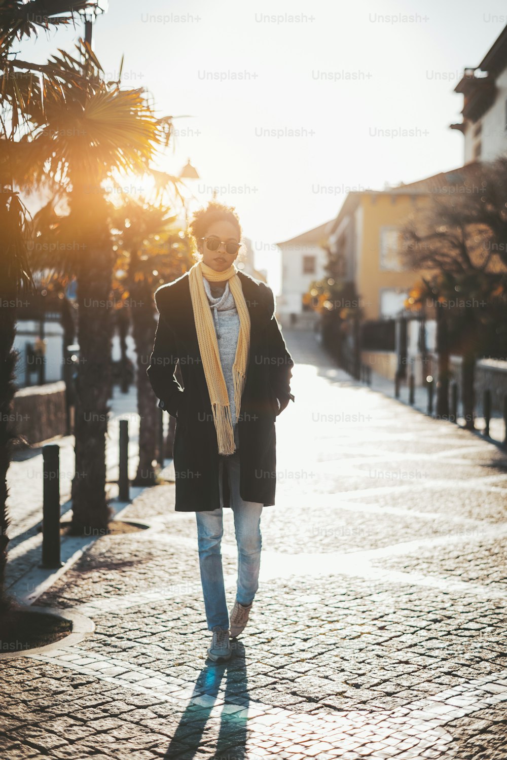 Vista vertical de una joven turista hipster afroamericana con un cálido abrigo de entretiempo y una bufanda iluminada desde atrás caminando por una calle estrecha con pavimento de piedra, con palmeras a la izquierda