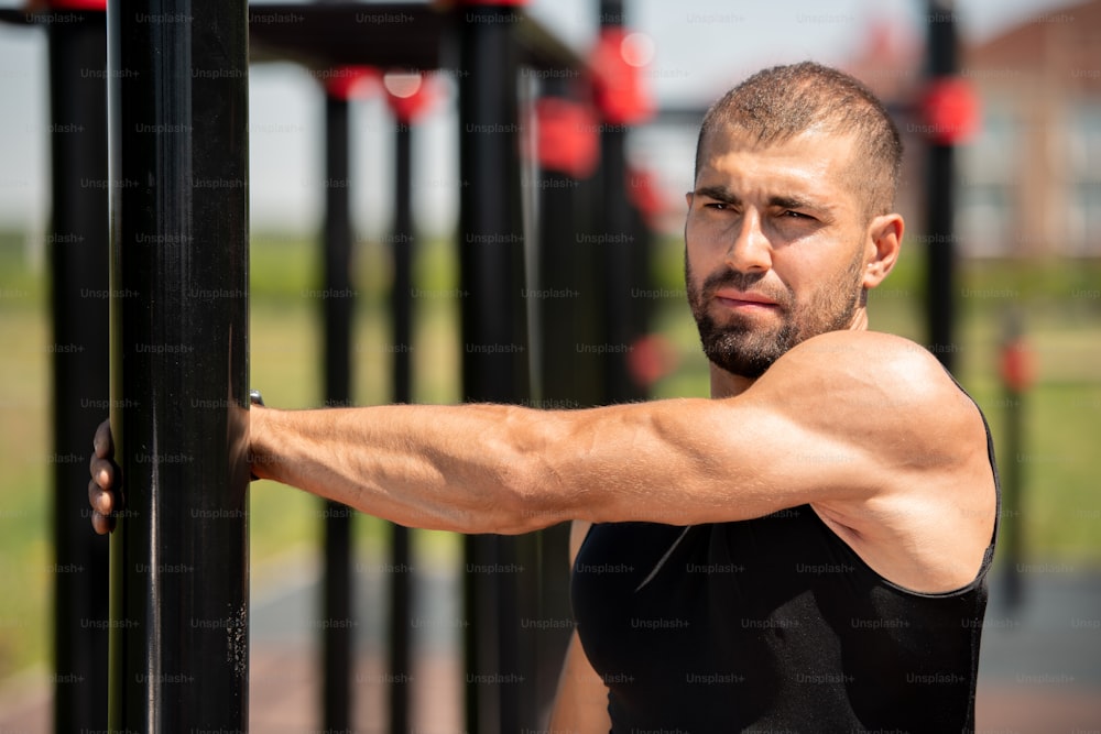 Muscular guy in black vest holding by sports bar while stretching arm during workout on sports facilities outdoors