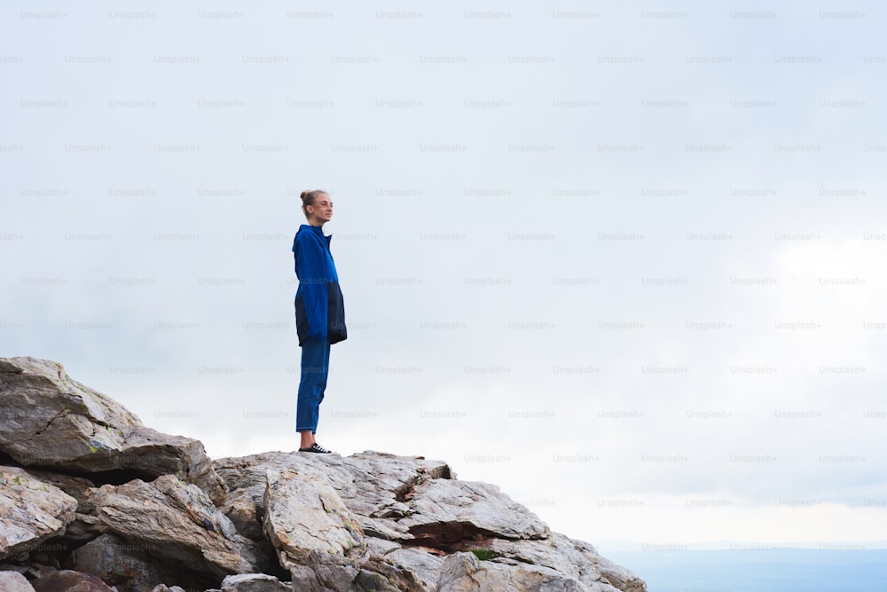 Woman standing on the top of the mountain after climbing, she's wearing blue jacket, against gloomy sky