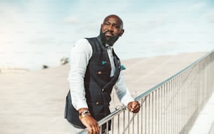 Portrait of a serious noble bald bearded African man in an elegant vest standing outdoors with the spectacles in a hand, holding a chromium-plated bridge fence and squinting looking aside