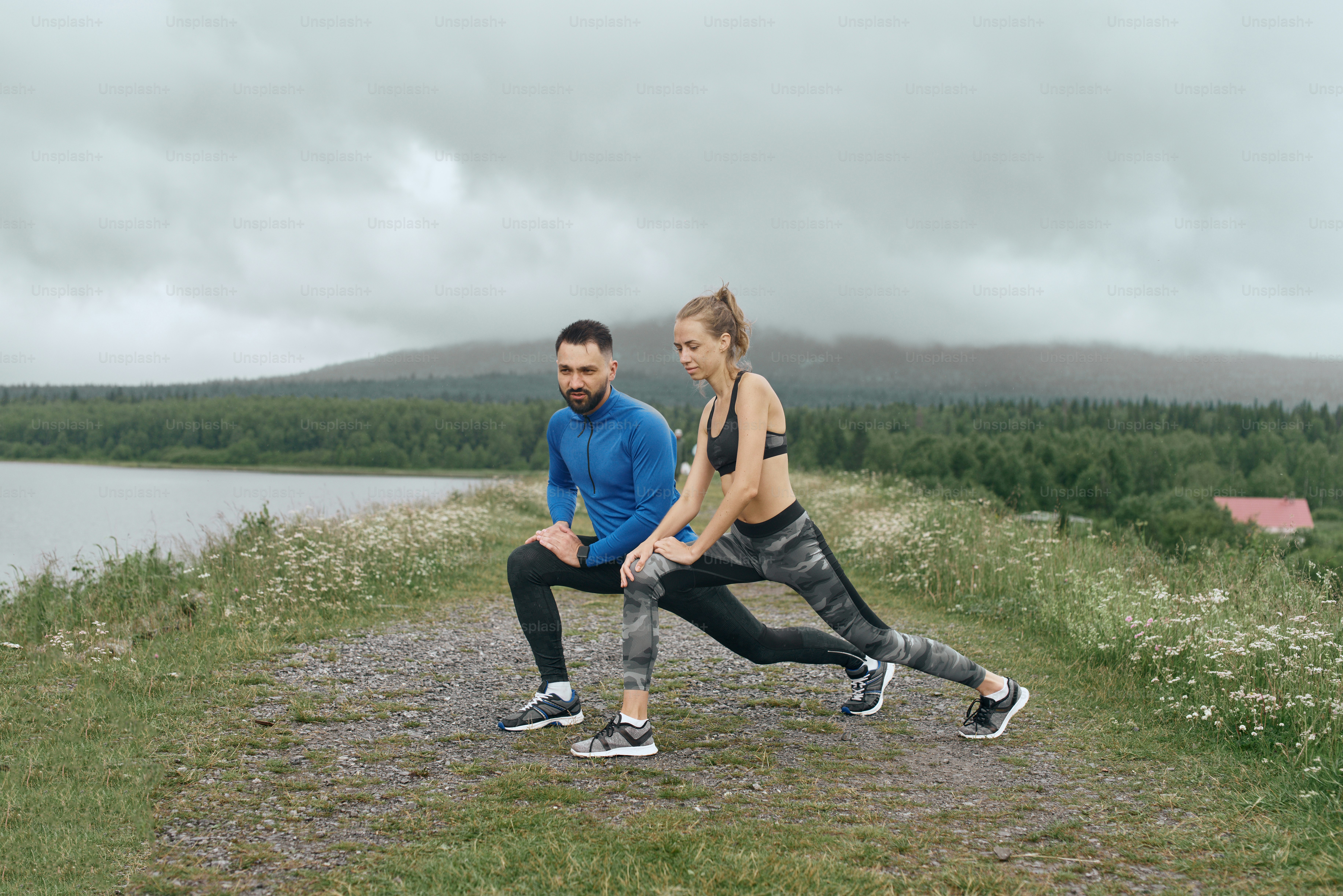 Two mid age man and woman exercising outdoor, warming up arm muscules, in summer, on gloomy day, standing at the road with scenic view, doing warm up stretching, wearing blue jacket, he has a beard