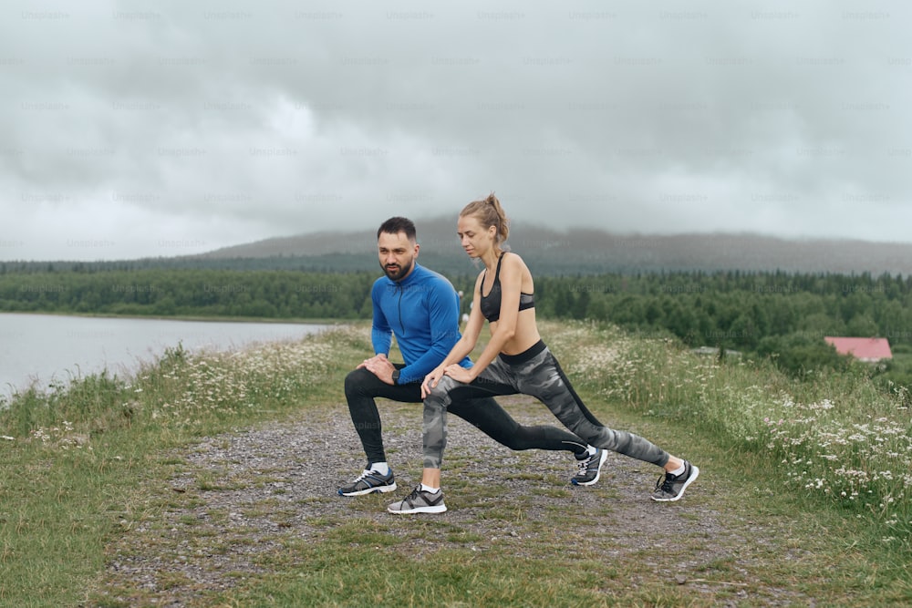 Two mid age man and woman exercising outdoor, warming up arm muscules, in summer, on gloomy day, standing at the road with scenic view, doing warm up stretching, wearing blue jacket, he has a beard