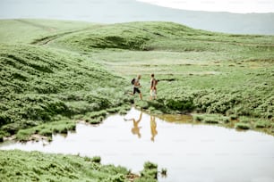 Man and woman hiking near the lake in the mountains, landscape view on the green meadow with lake during the sunny day