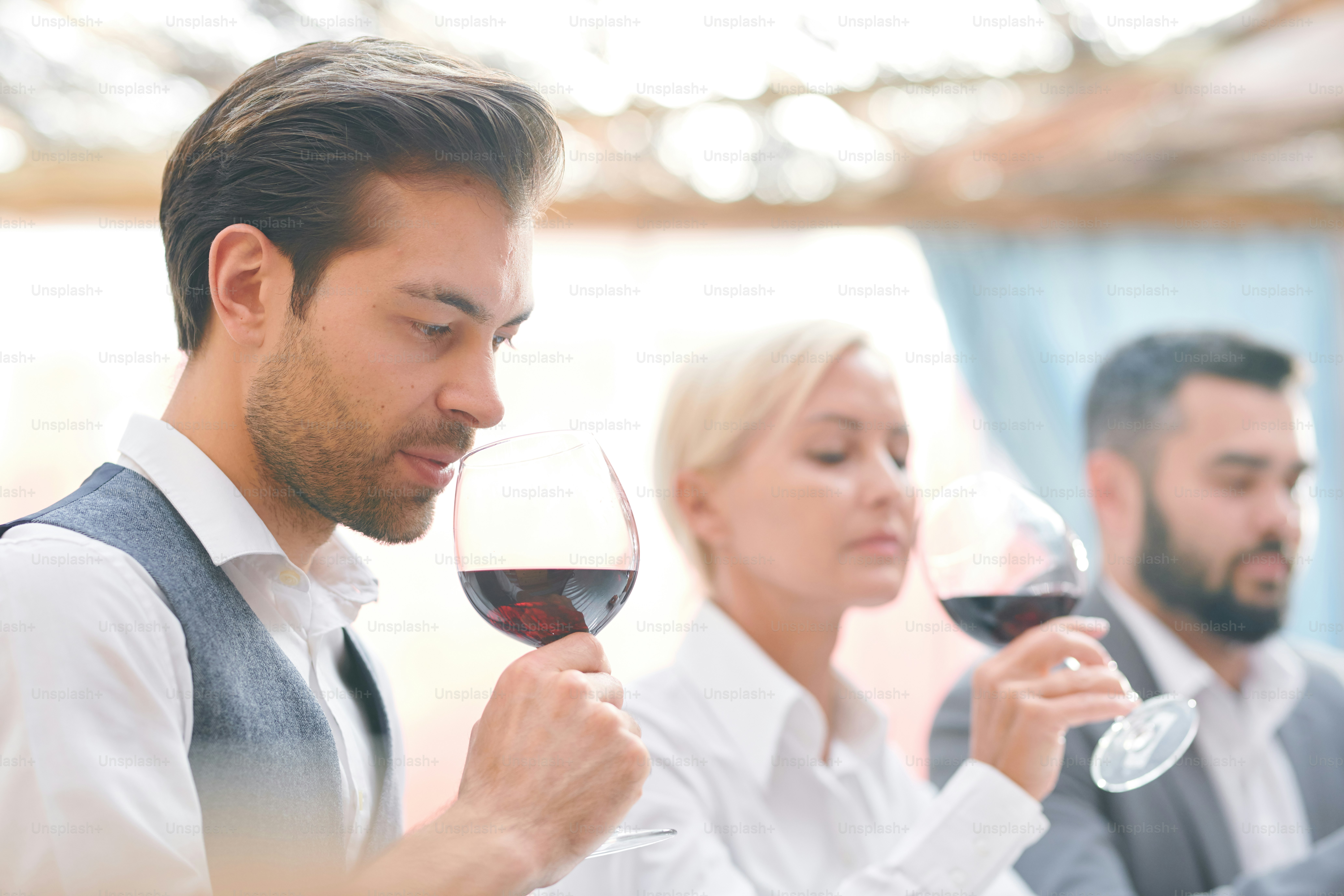 Sommelier masculin sérieux sentant le vin rouge du verre à vin tout en évaluant ses caractéristiques sur les antécédents de collègues