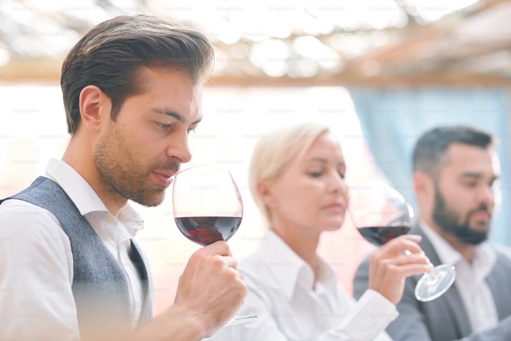 Serious male sommelier smelling red wine from wineglass while evaluating its characteristics on background of colleagues