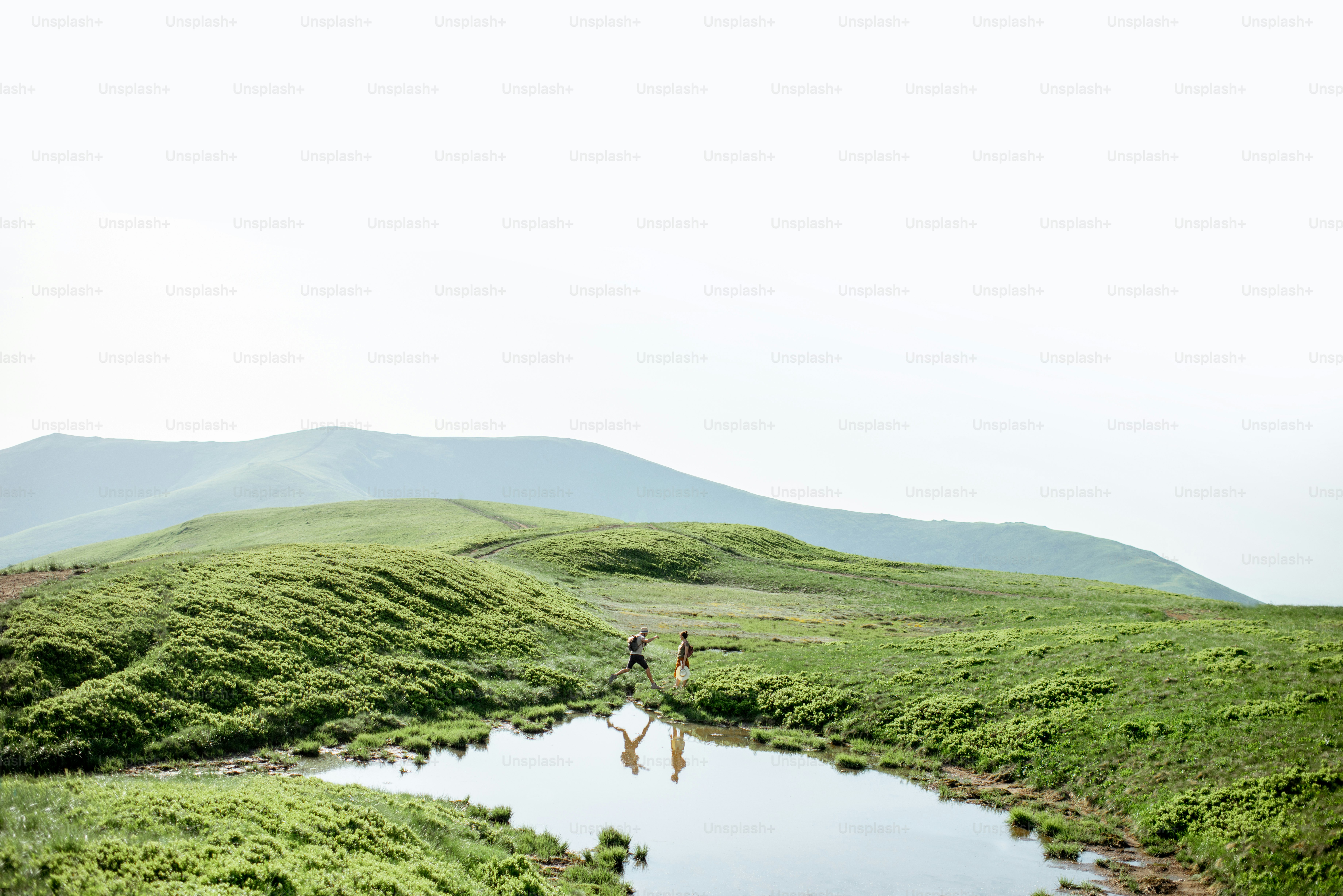 Man and woman hiking near the lake in the mountains, landscape view on the green meadow with lake during the sunny day