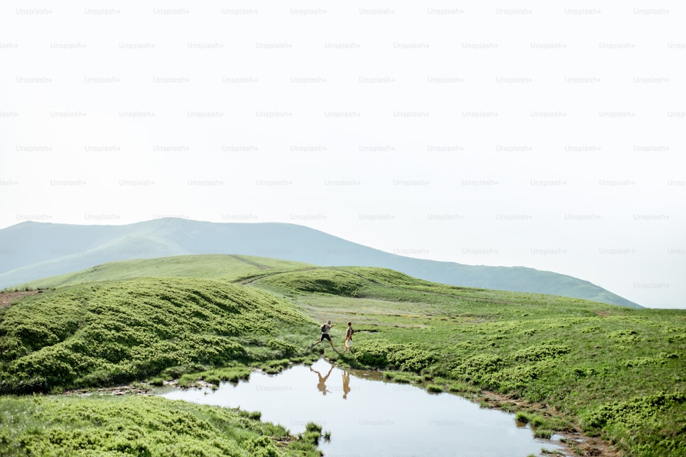 Man and woman hiking near the lake in the mountains, landscape view on the green meadow with lake during the sunny day
