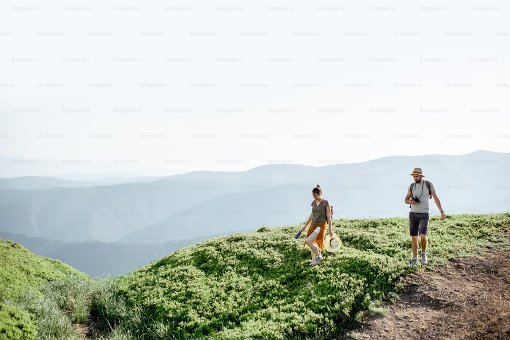 Beautiful couple walking with backpacks on the green meadow, while traveling high in the mountains during the summer time