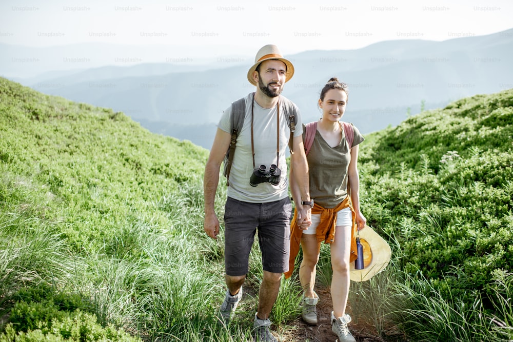 Beautiful couple walking with backpacks on the green valley, while traveling high in the mountains during the summer time